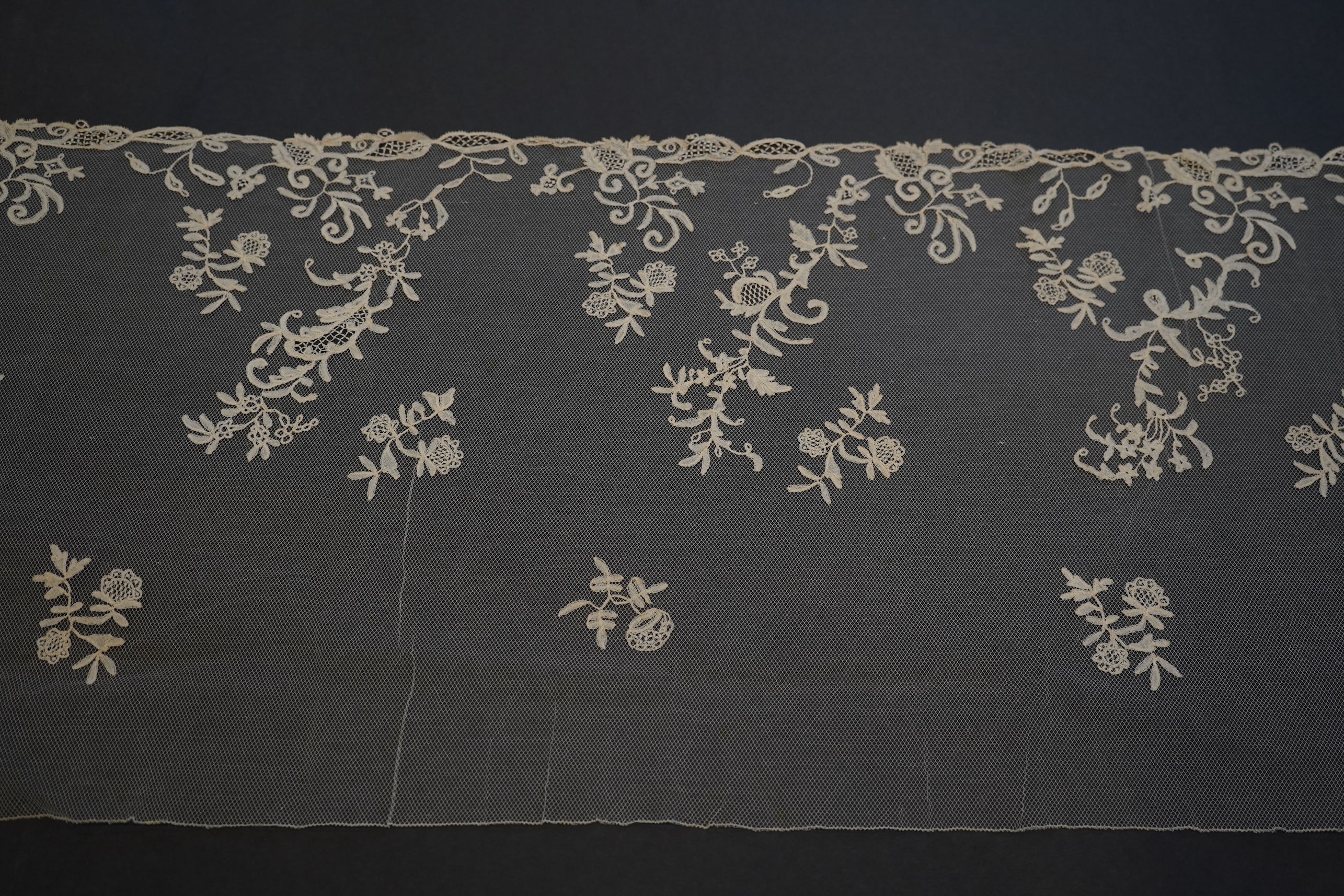 A 19th century five yard flounce of Brussels bobbin appliqué lace applied onto fine machine net, worked in a trailing floral design with an ornate border and sprig motifs, 457.5 cm long x 36cm deep. Condition - possibly